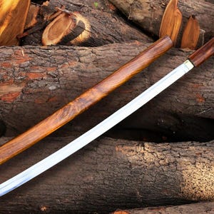 Könnte beinhalten: Ein japanisches Katana-Schwert mit einer Holzscheide und einem Griff, das auf einem Stapel Holzstämme liegt. Die Klinge ist silberfarben, und die Scheide und der Griff sind in dunklem Braun gehalten. Das Schwert ist teilweise gezogen.