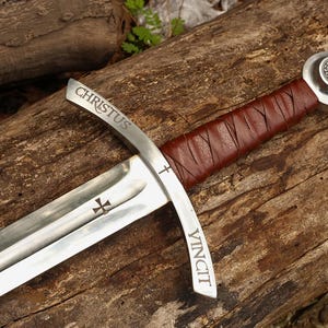 May include: A medieval-style sword with a silver blade and a brown leather-wrapped handle. The blade has the words "CHRISTUS VINCIT" etched on it, along with cross symbols. The sword rests on a weathered wooden surface.