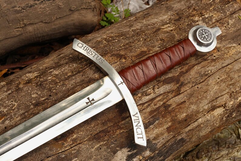 May include: A medieval-style sword with a silver blade and a brown leather-wrapped handle. The blade has the words "CHRISTUS VINCIT" etched on it, along with cross symbols. The sword rests on a weathered wooden surface.