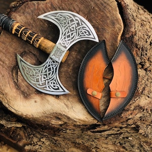 May include: A double-bladed axe with a silver head featuring intricate Celtic knot designs. The wooden handle has a dark, textured grip. A brown leather sheath with black trim and brass-colored fasteners is also present.