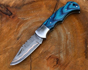 May include: A folding knife with a patterned blade and a vibrant blue wooden handle. The blade has a serrated edge and a copper-colored accent. The handle is secured with silver rivets.