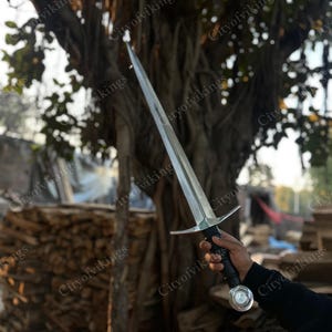 May include: A long, silver sword with a crossguard and a black handle is held in a person's hand. The sword has a sharp blade and a decorative pommel. The background includes a tree and a pile of wood.