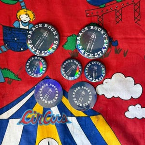 Könnte beinhalten: Eine Sammlung runder Buttons mit dem Text "GRACE ROCKY SAVE STARS" um eine Raketen-Grafik. Die Buttons haben einen holografischen Effekt und sind in verschiedenen Größen erhältlich. Das Wort "Circus" ist ebenfalls sichtbar.