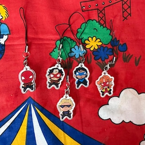 May include: Six superhero-themed acrylic charms with black lanyards and silver clasps. Each charm features a different cartoon character, with vibrant colours and bold outlines. The charms are displayed on a red patterned background.