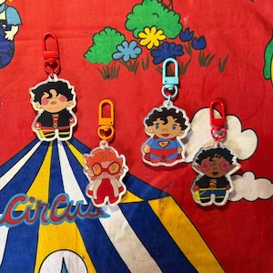 May include: Four cartoon character keyrings with red, orange, and blue clasps. The characters are wearing superhero-themed outfits. The background is a red fabric with a circus tent design.