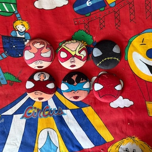 May include: Six hand-painted superhero-themed badges, each with a unique character design and vibrant colours. The badges are arranged on red fabric with a circus tent pattern. Each badge is round in shape.