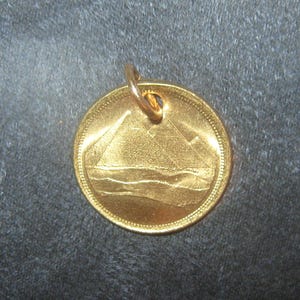 May include: A gold-colored pendant with a design of a pyramid and a wavy line, possibly representing a mountain and a river. The pendant has a small loop at the top for attaching a chain.