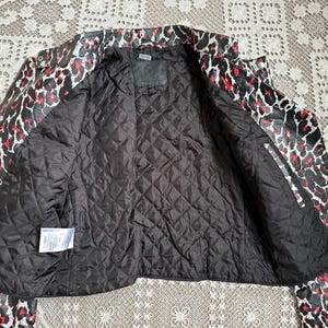 May include: A black, red, and white leopard print jacket with a quilted black lining. The jacket is open, revealing the interior lining and a small label. The collar and sleeves are visible, showcasing the bold pattern.