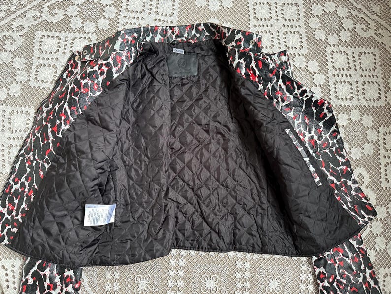 May include: A black, red, and white leopard print jacket with a quilted black lining. The jacket is open, revealing the interior lining and a small label. The collar and sleeves are visible, showcasing the bold pattern.