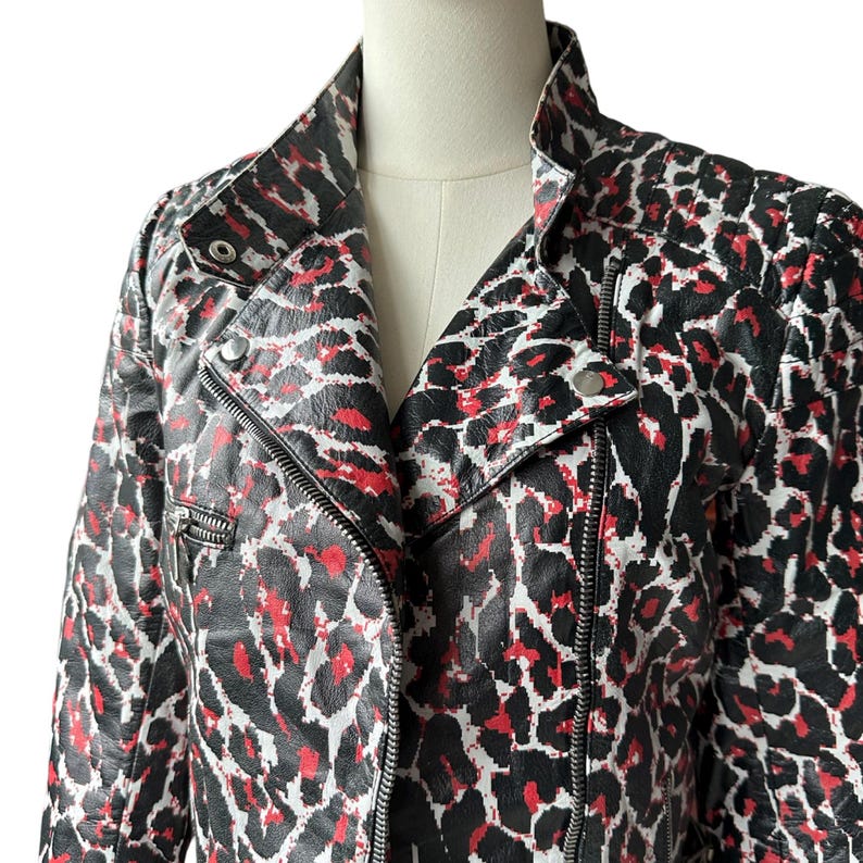 May include: A black, red, and white leopard print jacket with a classic biker style. The jacket features a notched collar, a diagonal zipper closure, and a small zippered pocket. The jacket is displayed on a mannequin.