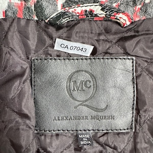 May include: Close-up of a black leather label with the Alexander McQueen logo and text. The label is stitched onto a quilted brown fabric. The top of the image shows a section of a jacket with a red, black, and white pattern.