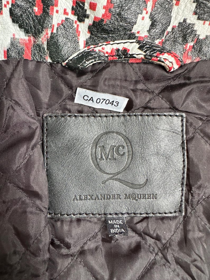 May include: Close-up of a black leather label with the Alexander McQueen logo and text. The label is stitched onto a quilted brown fabric. The top of the image shows a section of a jacket with a red, black, and white pattern.