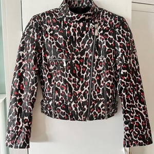May include: A black, white, and red leopard print jacket with a high collar and asymmetrical zipper closure. The jacket features two zippered pockets and silver hardware. The sleeves have zippers at the cuffs and adjustable buckles at the waist.