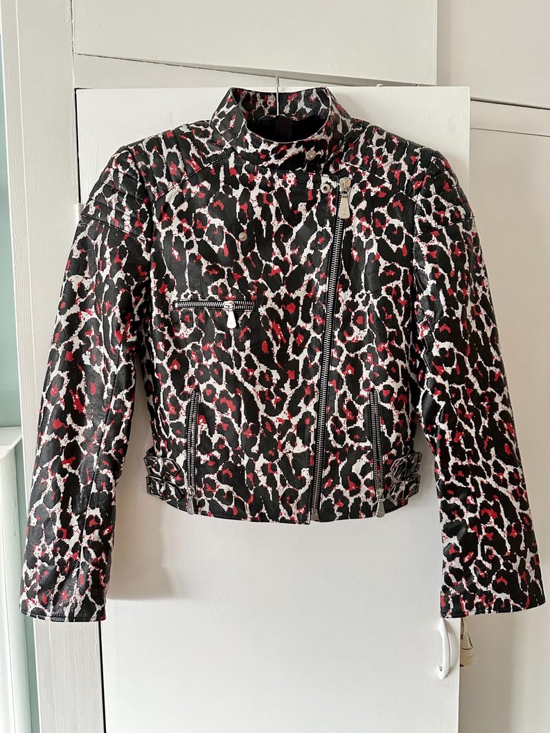 May include: A black, white, and red leopard print jacket with a high collar and asymmetrical zipper closure. The jacket features two zippered pockets and silver hardware. The sleeves have zippers at the cuffs and adjustable buckles at the waist.
