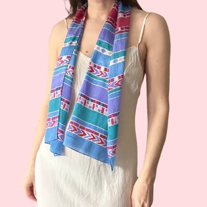 May include: A patterned scarf in shades of blue, purple, teal, and red. The scarf features a geometric design. It is draped around the neck. The scarf is rectangular and made of a lightweight material.