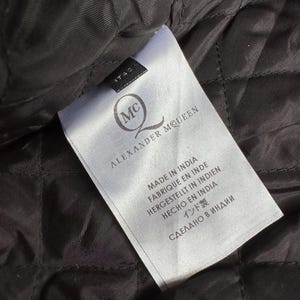 May include: Close-up of a white label with the text "ALEXANDER MCQUEEN" and the brand's logo. The label also includes text in multiple languages, including "MADE IN INDIA". The label is on a black quilted fabric.