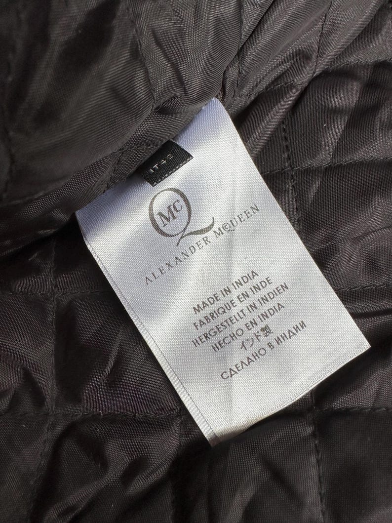 May include: Close-up of a white label with the text "ALEXANDER MCQUEEN" and the brand's logo. The label also includes text in multiple languages, including "MADE IN INDIA". The label is on a black quilted fabric.