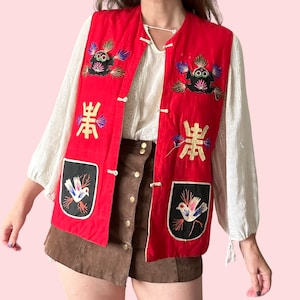 May include: A red vintage vest with embroidered details. The vest features black and white bird patches, Chinese characters, and frog-like designs. It has a button closure and is worn over a white blouse and brown shorts.