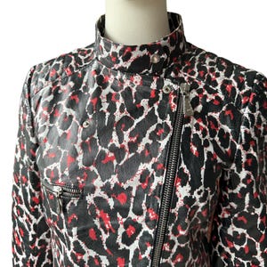 May include: A black, red, and white leopard print jacket with a high collar and a silver zipper. The jacket has a fitted silhouette and a quilted shoulder detail. The pattern is bold and eye-catching, with a mix of black, red, and white spots.