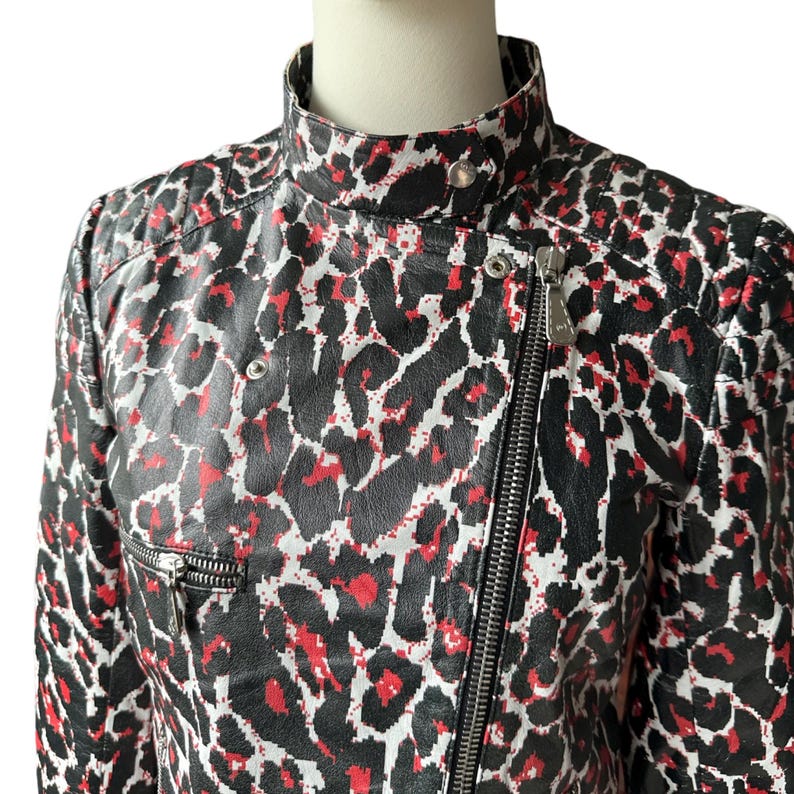 May include: A black, red, and white leopard print jacket with a high collar and a silver zipper. The jacket has a fitted silhouette and a quilted shoulder detail. The pattern is bold and eye-catching, with a mix of black, red, and white spots.
