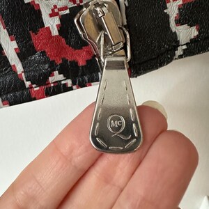 May include: Close-up of a silver zipper with a rectangular pull tab. The pull tab has the initials "MC" inside a circle. The zipper is attached to a black, red, and white patterned fabric.