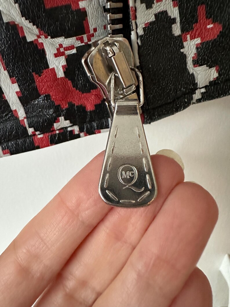 May include: Close-up of a silver zipper with a rectangular pull tab. The pull tab has the initials "MC" inside a circle. The zipper is attached to a black, red, and white patterned fabric.