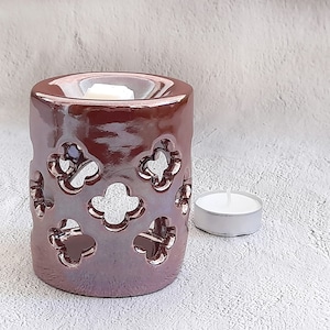 Puede incluir: Un quemador de aceite de cerámica marrón con un diseño decorativo de trébol. El quemador tiene una pequeña vela de té blanca a su lado.