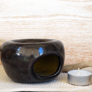 May include: A dark brown ceramic essential oil diffuser with a large opening and a small tea light candle. The diffuser has a glossy finish and sits on a white cloth. The background is a light wood.