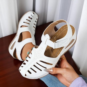 Könnte beinhalten: Weiße Sandalen mit Aussparungen und verstellbaren Riemen. Die Sandalen haben eine braune Innensohle und einen Klettverschluss. Das offene Design und die perforierten Details verleihen den bequemen Schuhen einen stilvollen Touch.