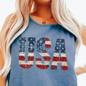 Könnte beinhalten: Hellblaues Tanktop mit dem Wort "USA" auf der Vorderseite, das im Design der amerikanischen Flagge abgebildet ist. Die Buchstaben sind rot, weiß und blau und haben einen Vintage-Look. Das Top ist ärmellos und aus weichem Material.