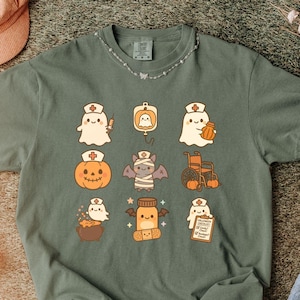 May include: Moss green t-shirt featuring a collection of cute Halloween-themed illustrations. The designs include ghosts, pumpkins, bats, and medicine bottles, all wearing nurse hats. The shirt has a crew neck and short sleeves.