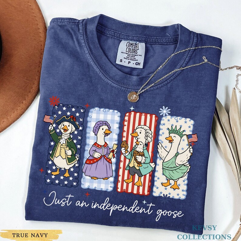 Pu&ograve; includere: T-shirt blu navy con una grafica di quattro oche vestite con abiti patriottici. La frase "Just an independent goose" &egrave; stampata sotto. Una collana d'oro &egrave; drappeggiata sulla maglietta.