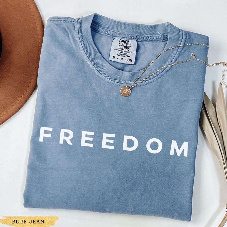 May include: A blue jean colored t-shirt with the word "FREEDOM" printed in white. A gold necklace with a pendant is draped over the shirt. A brown hat is visible in the upper left corner.