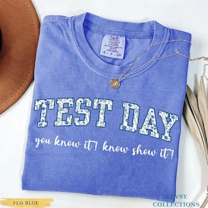 Könnte beinhalten: Ein blaues Comfort Colors T-Shirt mit den Worten "TEST DAY" in einem floralen Design. Darunter steht der Satz "you know it! know show it!" Eine goldene Halskette mit Anhänger befindet sich auf dem Shirt.
