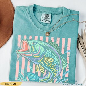 Op de afbeelding: Een zeegroen t-shirt met korte mouwen en een afbeelding van een vis met een open mond. Het ontwerp bevat verticale roze strepen en een verweerde textuur. Het label in de nek vermeldt Comfort Colors, S P CH, 100% katoen, made in Honduras.