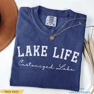 May include: A navy blue t-shirt with the words "LAKE LIFE" in white block letters and "Customized Lake" in a script font. A gold necklace with a pendant rests on the shirt. The shirt is labeled "TRUE NAVY".
