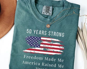 May include: Blue spruce t-shirt with the text "50 Years Strong" and an American flag design. The shirt also features the phrases "Freedom Made Me" and "America Raised Me" with "Since '75" below. A gold necklace is draped across the shirt.
