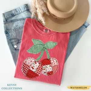 May include: A pink t-shirt with a cherry applique, green gingham leaves, and patchwork cherries in red and white patterns. The shirt is paired with light blue jeans and a tan hat. The text "KEVSY COLLECTIONS" and "WATERMELON" are visible.