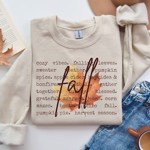 May include: A beige sweatshirt with the word "fall" in a script font and a leaf graphic. The sweatshirt has a list of fall-themed words such as "cozy vibes," "pumpkin," and "harvest season" printed on it.