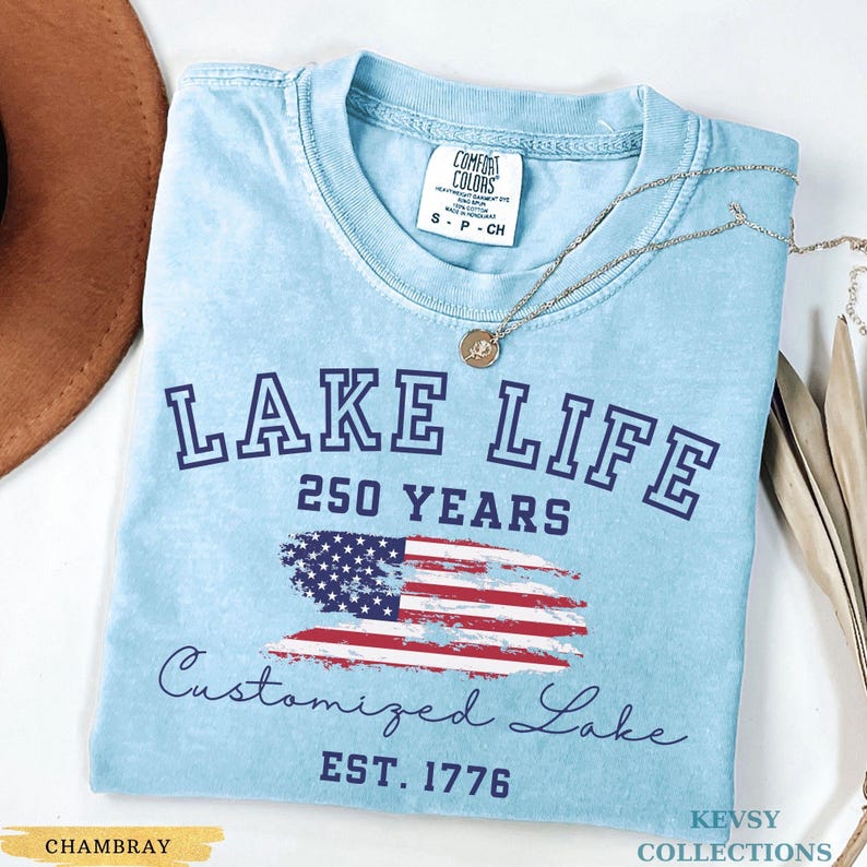Puede incluir: Camiseta azul claro con las palabras "LAKE LIFE" y "250 YEARS" en azul marino. Un gr&aacute;fico de la bandera estadounidense est&aacute; debajo del texto. La camiseta tambi&eacute;n tiene las palabras "Customized Lake" y "EST. 1776". Un collar plateado est&aacute; sobre la camiseta.