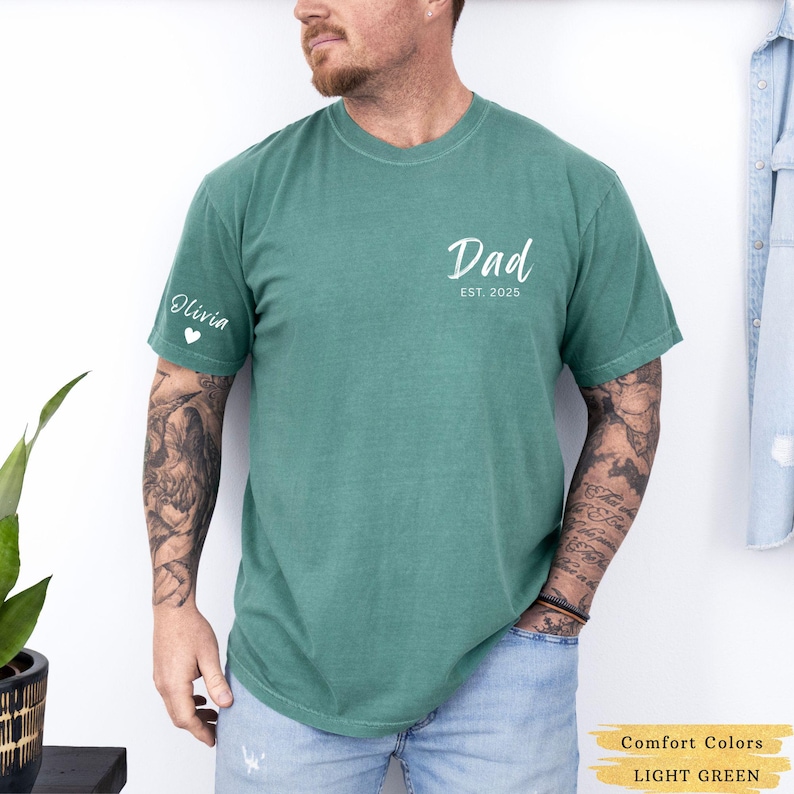 Może przedstawiać: Jasnozielony t-shirt z napisem "Dad Est. 2025" z przodu i napisem "Olivia" z sercem na lewym rękawie.