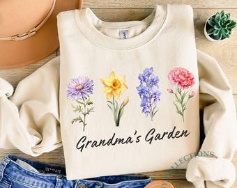 May include: Cream-colored sweatshirt featuring a floral design with the text "Grandma's Garden." The design includes watercolor illustrations of purple, yellow, blue, and pink flowers. The sweatshirt is paired with denim jeans and a brown hat.