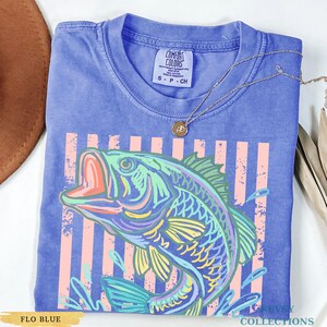 Op de afbeelding: Een periwinkle blauw t-shirt met korte mouwen en een afbeelding van een springende vis met neon-groene, gele en roze omlijningen. Het ontwerp bevat verticale lichtroze strepen achter de vis. Een Comfort Colors label is zichtbaar bij de nek.