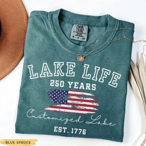 Puede incluir: Camiseta azul abeto con las palabras "LAKE LIFE 250 YEARS" en letras blancas. Debajo, un dise&ntilde;o de la bandera estadounidense y las palabras "Customized Lake EST. 1776". Un collar dorado est&aacute; sobre la camiseta.