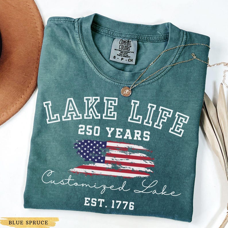 Puede incluir: Camiseta azul abeto con las palabras "LAKE LIFE 250 YEARS" en letras blancas. Debajo, un dise&ntilde;o de la bandera estadounidense y las palabras "Customized Lake EST. 1776". Un collar dorado est&aacute; sobre la camiseta.