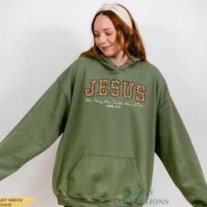 May include: A military green hoodie with a front pocket and a hood. The word "JESUS" is printed across the chest in a leopard print design, with the text "the Way the Truth the Life" and "JOHN 14:6" below it. The hoodie is a comfortable, casual style.