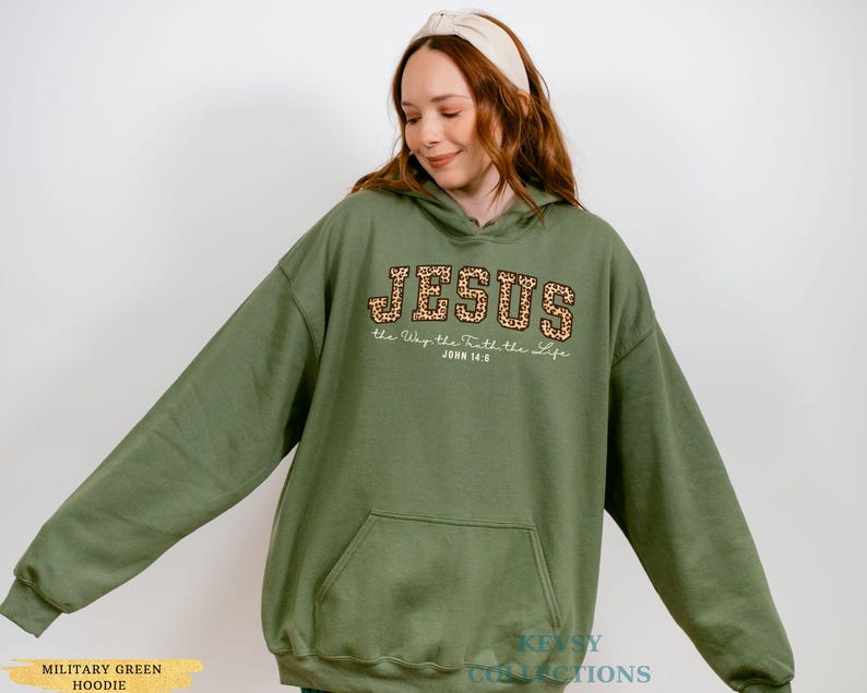 May include: A military green hoodie with a front pocket and a hood. The word "JESUS" is printed across the chest in a leopard print design, with the text "the Way the Truth the Life" and "JOHN 14:6" below it. The hoodie is a comfortable, casual style.