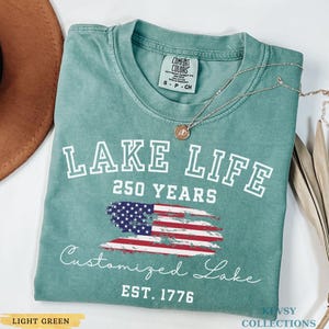 Puede incluir: Camiseta verde claro con las palabras "LAKE LIFE" y "250 YEARS" en blanco. Debajo, una bandera americana estilizada y las palabras "Customized Lake" y "EST. 1776". Un collar plateado con un colgante est&aacute; sobre la camiseta.