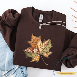 May include: A dark chocolate-coloured sweatshirt with a detailed, patchwork-style leaf design. The leaf features various patterns and colours, including orange, green, and brown. A silver necklace with a small pendant is draped on the sweatshirt. The text "DARK CHOCOLATE" is visible.