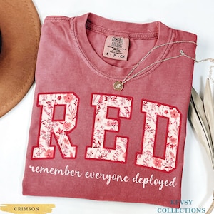 May include: Crimson-colored t-shirt with the word "RED" in large, floral-filled letters. The text "remember everyone deployed" is below the letters. A silver necklace with a pendant is on the shirt.